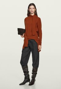 Frau in einem rost-orangefarbenen Pullover mit Schal-Details, dunklen, weiten Hosen und schwarzen, lässigen Stiefeln, die eine schwarze Clutch vor einem schlichten Hintergrund hält.