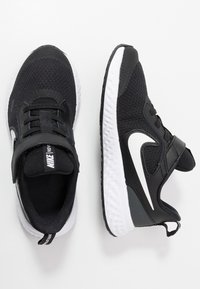 Zapatillas Nike negras con parte superior de malla, acentos de cuero, correa ajustable y suelas de goma blanca con superficie texturizada y logotipo.