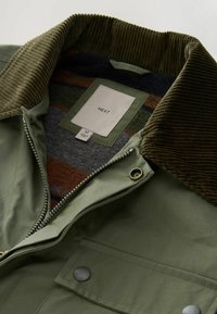 Olivgrüne Jacke mit einem Cordkragen, Reißverschluss und innenseitiger gestreifter Fütterung in Grau, Orange und Grün. Sie verfügt über eine Fronttasche.