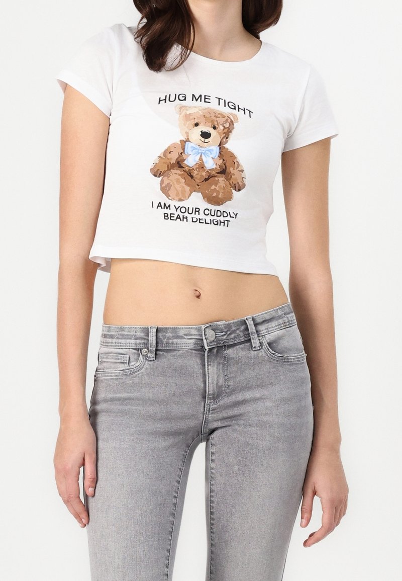 Camiseta corta blanca con un gráfico de un oso de peluche marrón que lleva un lazo azul. El texto dice: "ABRÁZAME FUERTE, SOY TU DELICIA DE OSITO ACOGEDOR".