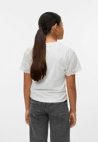 Camiseta blanca con detalles de encaje en las mangas cortas, combinada con jeans gris oscuro. La parte superior tiene un corte holgado y un diseño simple.