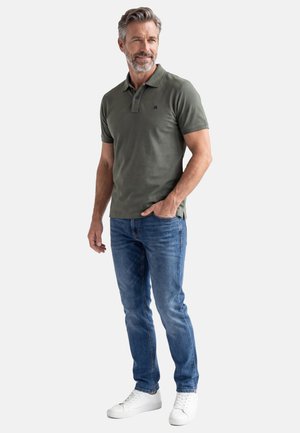 Uomo di mezza età con capelli e barba grigi, che indossa una polo verde oliva, jeans blu e scarpe da ginnastica bianche, in piedi con una mano in tasca.