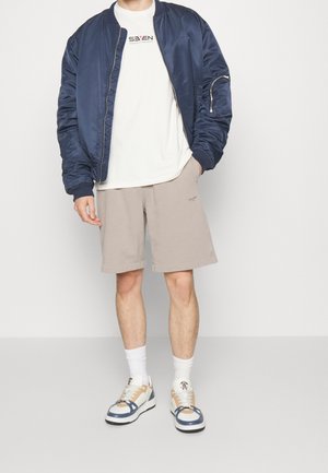 Marineblauwe bomberjack, wit T-shirt met afbeelding, beige korte broek, witte sokken en beige sneakers met blauwe accenten. Casual outfit getoond.