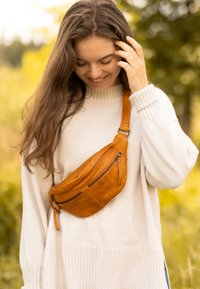 Smeđa kožna fasin torbica s zakrivljenim oblikom, dva patentna zatvarača i podesivim remenom, nošena preko krem pletenog džempera.