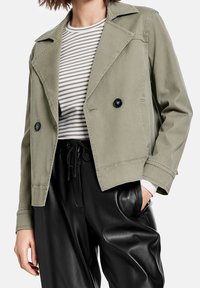 Krótka kurtka w oliwkowym kolorze z dwurzędowym zapięciem, czarnymi guzikami i miękką fakturą, zestawiona z koszulą w paski i czarnymi spodniami.