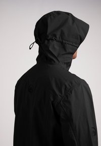 Mand iført en sort vandtæt jakke med hætten oppe, vist fra baggrunden mod en ensfarvet lys baggrund.