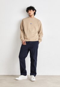 Sweatshirt beige a collo interno con un piccolo logo blu, abbinato a pantaloni navy e sneakers bianche. Tessuto morbido, vestibilità comoda, design minimale.