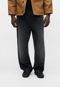 Άνδρας που φοράει μαύρα τζιν με φαρδιά μπατζάκια και εμπρόσθιες τσέπες, μαύρα παπούτσια και καφέ μπουφάν, μπροστά σε απλό φωτεινό φόντο.