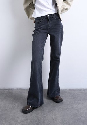 Person iført sorte højtaljede flared jeans, hvid skjorte, beige jakke og mørkebrune platåsko stående på et betongulv.