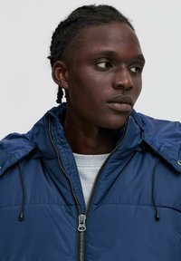 Tumši zils pūkains jaka ar augstu apkakli, rāvējslēdzēja aizdari, savilcēju un gludu tekstūru. Uzvilkta virs gaiši pelēka krekla.