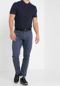 Homme portant un polo rayé bleu marine, un pantalon slim bleu, une ceinture noire, des chaussettes blanches et des chaussures de sport noires, debout devant un fond blanc.
