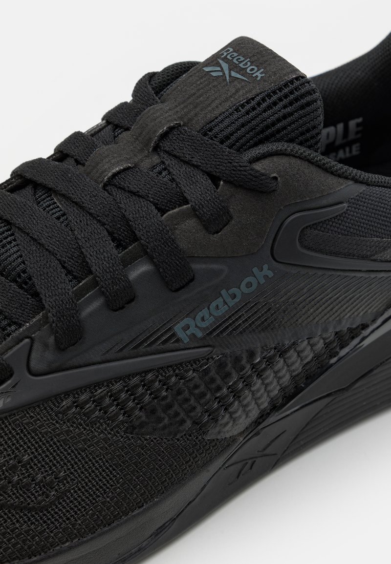 Acercamiento a una zapatilla negra de Reebok con malla texturizada, paneles de ante, lengüeta con marca y cierre con cordones.