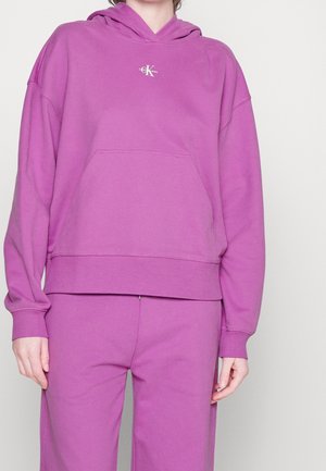 Felpa con cappuccio in cotone viola con tasca frontale, polsini e orlo a coste. Presenta un piccolo logo sul petto. Abbinata a pantaloni viola coordinati.