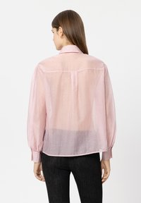 Woman with straight brown hair wearing a sheer pink long-sleeve blouse with white dots and black pants, facing away against a white background.