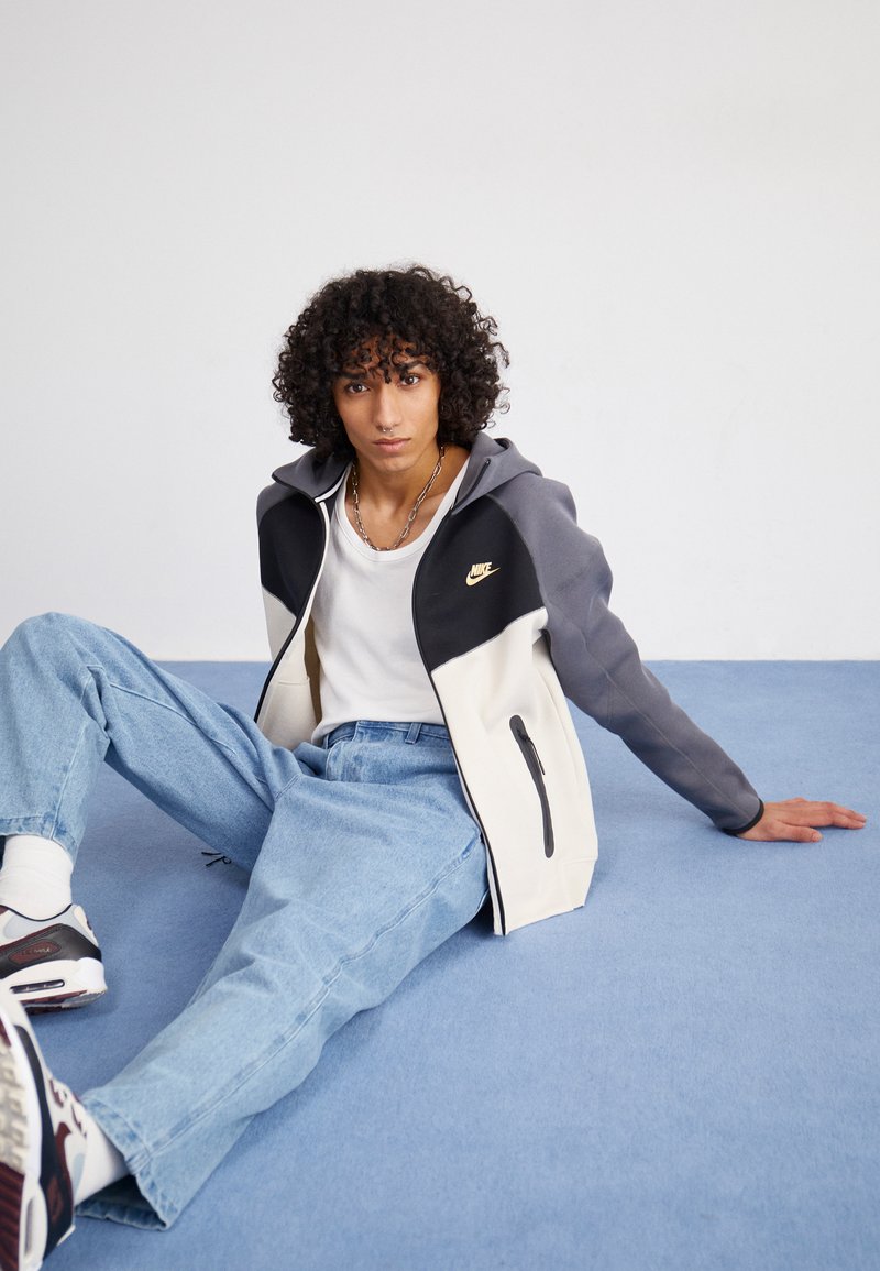 Felpa con zip Nike con un design a blocchi di colore nero, grigio e crema, caratterizzata da un logo dorato, cappuccio regolabile e tasche laterali con zip.