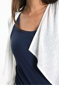 Cardigan léger blanc superposé sur un débardeur côtelé marine. Le cardigan présente un design texturé avec une ouverture à l'avant et un tissu drapé.