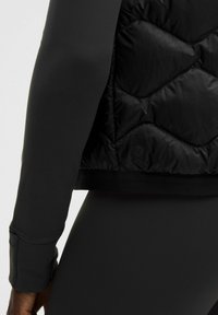 Musta toppaliivi, jossa tikattu kuvio, sileä pinta ja joustavat hihansuut. Kangas vaikuttaa kevyeltä ja vettä hylkivältä.