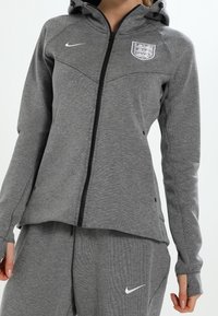 Sudadera con cremallera gris y cuello alto, que presenta el logotipo de Nike y el emblema de Inglaterra. Fabricada con un tejido texturizado, cuenta con mangas largas y bolsillos laterales.