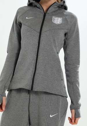 Grå lynlås hoodie med høj krave, featuring et Nike-logo og det engelske våbenskjold. Fremstillet af tekstureret stof, med lange ærmer og sidelommer.