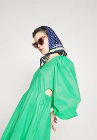 Vestido verde oversized com mangas bufantes e cintura ajustável. Combinando com um lenço azul marinho com bolinhas e óculos de sol cor-de-rosa, exibindo cores vibrantes.