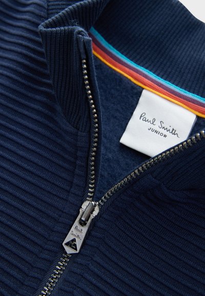 Tumši zils ribsētā rāvējslēdzēja jaka ar krāsainu svītrainu apkakli. Aprīkota ar metāla rāvējslēdzēju ar trīsstūra piekariņu un etiķeti ar uzrakstu "Paul Smith Junior."