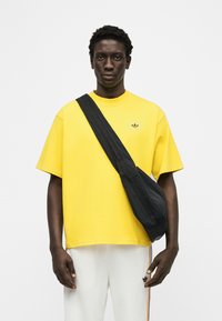 Άνδρας που φοράει φωτεινό κίτρινο πουκάμισο Adidas, λευκό παντελόνι Adidas με ρίγες και μαύρη τσάντα χιαστί, μπροστά σε απλό λευκό φόντο.