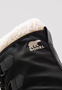 Czarne śniegowce z teksturowaną warstwą zewnętrzną, białym futrem polarnym i logo Sorel. Cechy obejmują dwie czarne taśmy oraz gładkie, trwałe wykończenie.