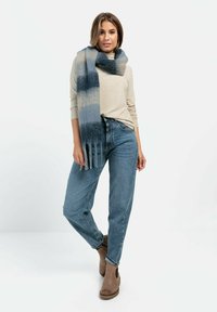 Wollmischungsschal in Blau- und Grautönen mit weicher Textur und Fransen, getragen über einem hellbeigen Oberteil und Jeans.