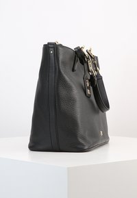 Svart läderhandväska med en texturerad utsida, guldpläterade detaljer, dubbla handtag och en avtagbar rem. Sido vy som visar form och design.