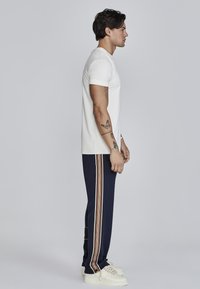 Camiseta blanca de manga corta combinada con pantalones de chándal azul marino con rayas verticales en naranja, blanco y marrón. Zapatillas blancas completan el look.