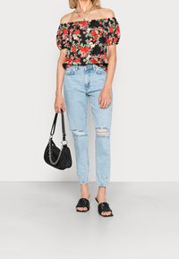 Femme portant un haut fleuri noir à épaules dénudées, un jean déchiré bleu clair, des sandales noires en toile à enfiler, tenant un sac noir avec une chaîne en argent.