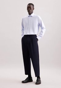 Camicia azzurro chiaro con colletto strutturato, abbinata a pantaloni plissettati blu navy. Scarpe nere stringate completano il look.