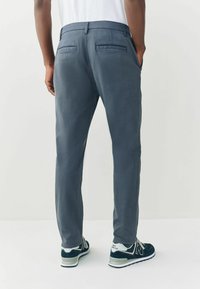 Férfi szürke nadrág sima textúrával, két hátsó zsebbel és testre szabott szabással, sötét atlétikai cipővel kiegészítve.