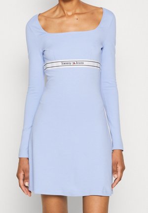 Jasnoniebieska sukienka z długim rękawem i kwadratowym dekoltem, ozdobiona centralnym białym pasem z napisem "Tommy Jeans" w czarnych literach.