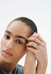 Anillo de oro con una piedra verde, llevado en una mano que descansa cerca de la cara de una persona con piel suave y cabello peinado hacia atrás.