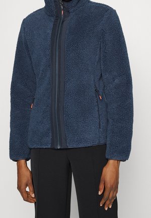 Persoon draagt een marineblauwe, gestructureerde fleecejas met rits en zwarte broek, staand met de armen ontspannen langs het lichaam.