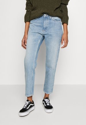 Person iført lyseblå højttaljede jeans, sorte og hvide sneakers samt en mørkegrøn strikket trøje stående på en grå gulv.