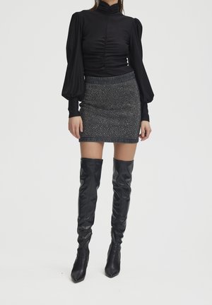 Kobieta w czarnej marszczonej bluzce z wysokim kołnierzem i bufkami, czarno-białej mini spódnicy w plamki oraz czarnych, sięgających uda kozakach.