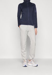 Chaqueta de forro polar con cierre en el frente en color azul marino y cuello alto, pantalones de sudadera en gris claro y zapatillas de running grises con acentos azules. Cuenta con bolsillos laterales.