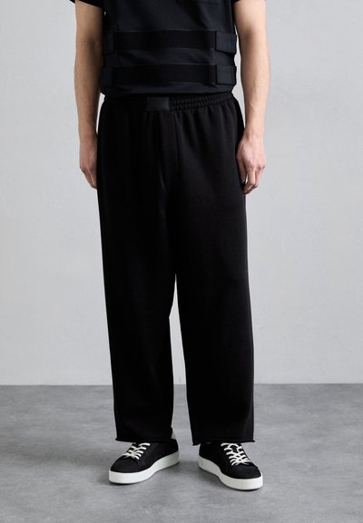 Pantaloni neri a vestibilità comoda con vita elastica, abbinati a un top nero con spalline orizzontali e sneakers nere con suola bianca.