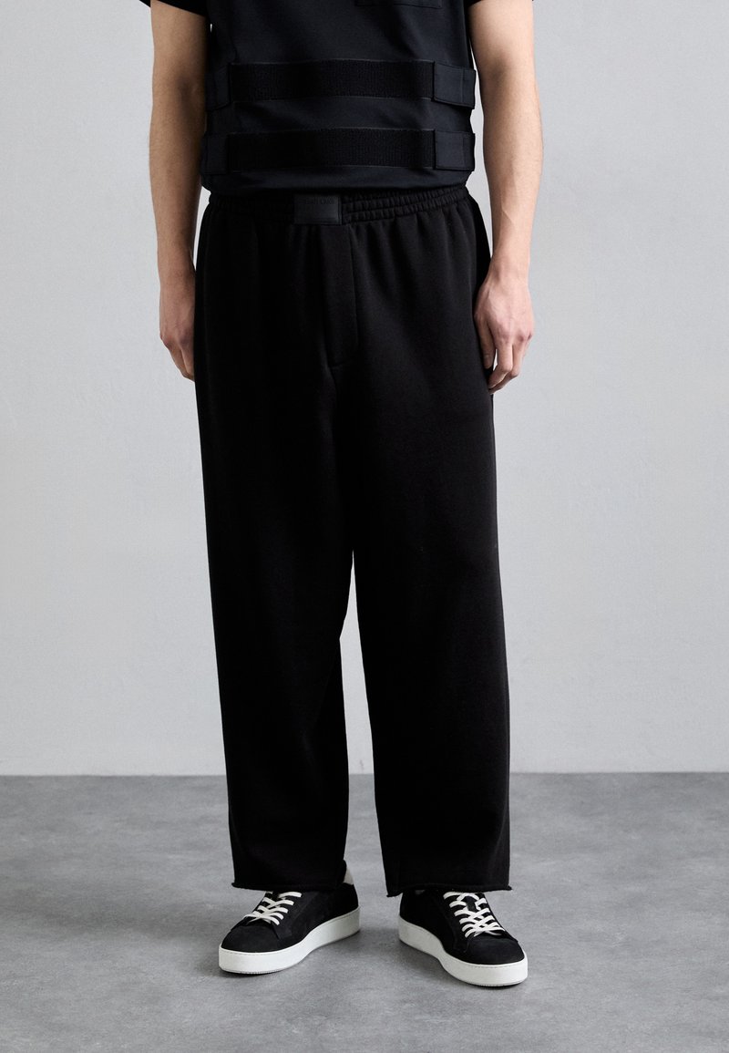 Schwarze lockere Hose mit elastischem Bund, kombiniert mit einem schwarzen Oberteil mit horizontalen Trägern und schwarzen Sneakers mit weißen Sohlen.