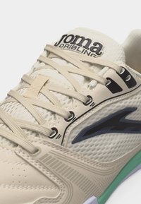 Beige ja rohelise spordijalatsi lähedalt, võrkse ülaosaga, beežide paeltega, musta logoga ja Joma Driblinguga bränditud keelega.