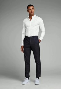 Weißes Hemd mit langen Ärmeln, dunklen Hosen und weißen Turnschuhen. Das Outfit hat ein figurbetontes Design und glatte Texturen.