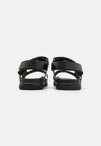 Black sandals with adjustable ankle straps, flat soles, and a minimalist design. Made of leather with a matte texture and simple hardware.