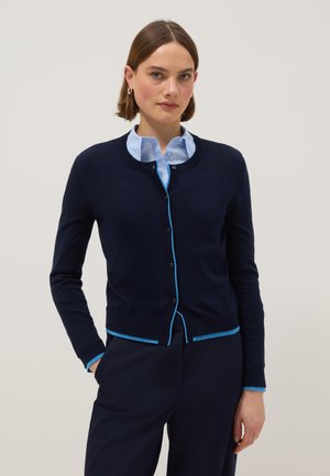 Giovane donna che indossa un cardigan blu navy con rifiniture azzurro chiaro, sopra una camicia con colletto azzurro chiaro, in piedi con una mano in tasca.