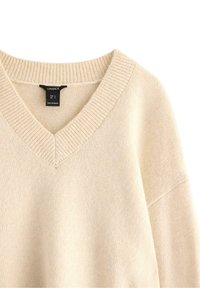 Beige svetr s výstřihem do V a žebrovaným límcem a rukávy, který vykazuje jemnou strukturu. Viditelná etiketa se značkou a informacemi o velikosti.