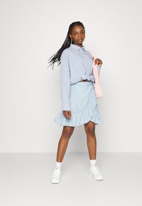 Camisa azul clara de botões atada na cintura, combinada com uma saia rodada azul clara com uma bainha com babados. Ténis brancos e uma bolsa rosa.