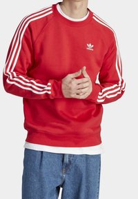 Felpa rossa con strisce diagonali bianche sulle maniche, collo e orlo a coste, con un piccolo logo bianco sul petto. Vestibilità casual.