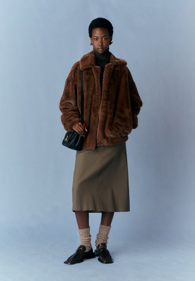 Braune Kunstpelzjacke mit breitem Kragen, kombiniert mit einem dunklen Rollkragenpullover, einem olivgrünen knielangen Rock, beigen Socken und dunklen Loafers. Schwarze Handtasche.