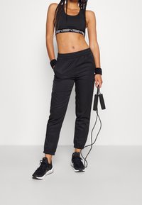 Czarne sportowe komplety składające się z dopasowanego cropped topu z paskiem z logo oraz zwężanych spodni dresowych. Akcesoria obejmują opaski na nadgarstki i skakankę.